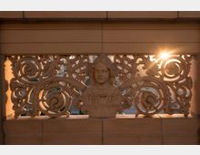 Verzierte Steinskulptur mit floralen Mustern und einem Relief eines weiblichen Gesichts in der Mitte, durchbrochen und teilweise von Sonnenlicht beleuchtet, das durch die Ornamente hindurchscheint. Im Hintergrund sind verschwommen Gebäude und eine Straße erkennbar.