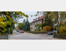 Wohnstraße mit Kopfsteinpflaster und herbstlicher Begrünung, umgeben von Bäumen und Sträuchern. Auf der rechten Seite ein zweistöckiges Haus mit rotem Ziegeldach und Efeu bewachsener Fassade, daneben geparkte Autos und Straßenlaternen.