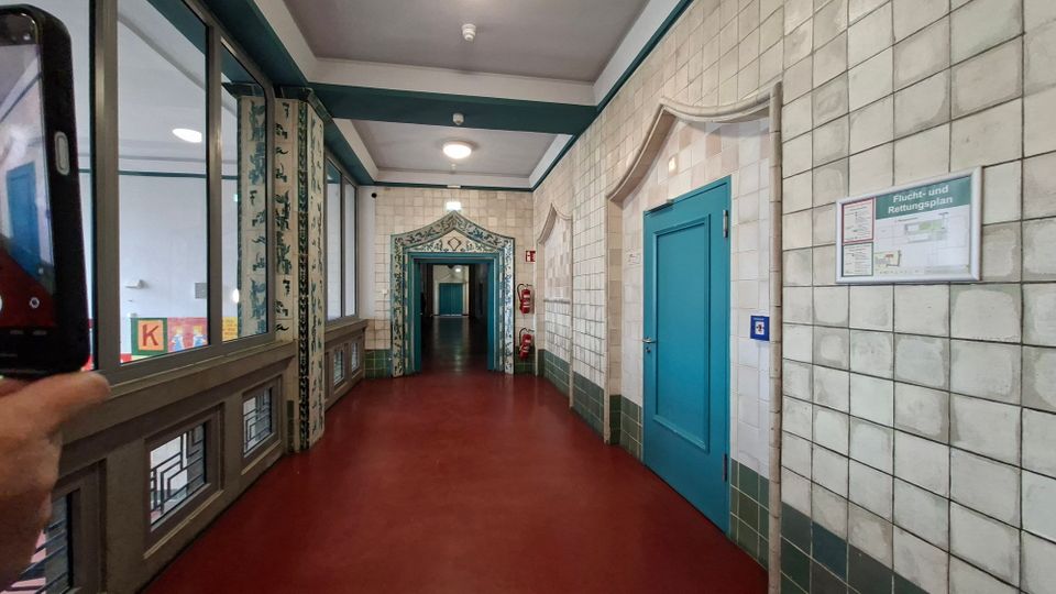 Langer Innenflur mit roten Bodenbelag, hellen Kachelwänden und türkisfarbenen Türen, beleuchtet durch Deckenlampen. Links verläuft eine Fensterfront mit dekorativen Glasfeldern, am Ende ist ein ornamental gerahmter Durchgang zu weiteren Fluren sichtbar.