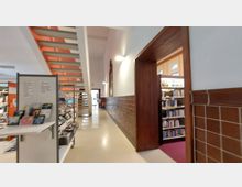 Innenansicht einer Bibliothek mit hohen Decken. Links befinden sich Regale mit Büchern und Zeitschriften, eine moderne Treppe aus Metall führt nach oben, und rechts öffnet sich ein Durchgang mit Holzrahmen zu einem weiteren Bereich mit Bücherregalen.