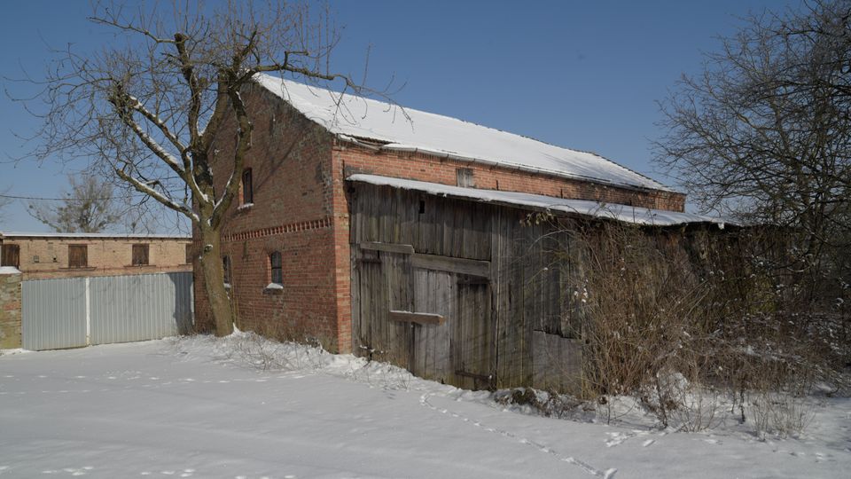 Ländlicher Hofbereich im Winter mit einem roten Backsteingebäude und einem angebauten Holzschuppen, dessen Dach mit Schnee bedeckt ist. Im Vordergrund liegt eine verschneite Fläche mit Fußspuren, umgeben von kahlen Bäumen und Sträuchern unter klarem blauem Himmel.
