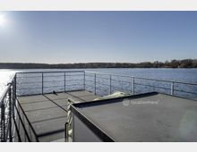 Eine Terrasse mit Metallgeländern auf einem Schiff oder einer Plattform, die über einen See hinausführt. Im Hintergrund ist eine ruhige Wasseroberfläche und eine bewaldete Uferlinie unter klarem, sonnigem Himmel zu sehen.