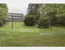 Weitläufige Grünfläche mit vereinzelten Gänseblümchen, umgeben von Bäumen und Sträuchern, im Hintergrund ein großes Gebäude mit Satteldach, mehreren Fenstern und einer Natursteinmauer. Links steht eine einfache Laterne, rechts schlängelt sich ein kleiner Pfad durch das Gras.