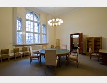 Ein Konferenzraum mit einem großen runden Holztisch, umgeben von gepolsterten Stühlen. Der Raum hat hohe Fenster mit Sprossen und eine zentrale Deckenleuchte, zusätzliche Holzmöbel wie Bücherregale und kleinere Tische stehen entlang der Wände.