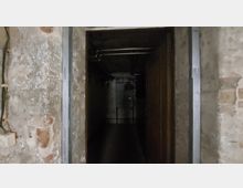 Ein schmaler Durchgang in einem Kellerraum mit unverputzten, teils abblätternden Wänden aus altem Mauerwerk. Im Hintergrund sind Rohre und metallische Konstruktionen an der Decke sichtbar.