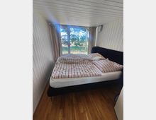 Schmales Schlafzimmer mit weißer Holzvertäfelung an Wänden und Decke, Holzboden und einem großen Bett mit gestreifter Bettwäsche. Am Kopfende befindet sich eine bodentiefe Fensterfront mit Vorhängen und Blick ins Grüne.