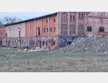 Altes Backsteingebäude mit gemischtem Mauerwerk aus Ziegel- und Natursteinen, davor eine Wiese mit weidenden Schafen und ein Hügel aus Steinen und Erde. Im Hintergrund sind vereinzelt kahle Bäume zu erkennen.