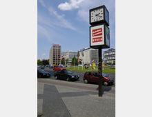 Straßenszene mit einer freistehenden Uhr und einem Werbeschild in der Nähe eines gepflasterten Gehwegs, dahinter mehrspurige Fahrbahn mit geparkten Autos und Bürogebäuden. Im Hintergrund ragen Flaggenmasten und ein modernes Gebäude mit dem Schriftzug „Eternit“ hervor.