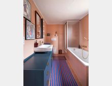 Ein Badezimmer mit aprikosenfarbenen Wandfliesen und einem braunen Fliesenboden. Auf der linken Seite steht ein dunkelblaues Waschbeckenmöbel mit einem weißen Waschbecken und goldfarbenen Armaturen, rechts ist eine Badewanne mit Glasduschabtrennung. Zwei gemusterte Teppiche in Blau und Rot bedecken den Boden, und gerahmte Kunstwerke dekorieren die Wände.