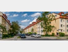 Wohnstraße mit gelblichen Mehrfamilienhäusern im Stil der 1930er-Jahre, die parallel zueinander verlaufen. Der Straßenrand ist mit parkenden Autos gesäumt, und dazwischen stehen Bäume und grüne Büsche; im Hintergrund ein blauer Himmel mit einigen Wolken.