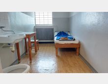 Schlicht eingerichtete Zelle mit weiß gestrichenen Wänden, einem kleinen Bett mit Bettzeug, einem Holztisch mit Hocker und einem Waschbecken. Tageslicht fällt durch ein vergittertes Fenster im Hintergrund.