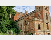 Eine verfallene, historische Backsteinvilla mit beschädigten Fenstern und bröckelndem Putz, umgeben von dichtem Grün. Der Bau weist gotisch inspirierte Details wie Zinnen auf und steht in einer natürlichen Umgebung mit Bäumen und hohem Gras.