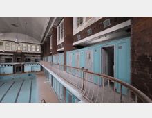Innenraum eines verlassenen Schwimmbades mit einem leeren Schwimmbecken im Vordergrund. Im Hintergrund sind türkisfarbene Umkleidekabinen und eine Galerie mit Geländer sichtbar, darüber hohe Fenster und eine gewölbte Decke.