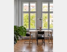 Helles Wohnzimmer oder Esszimmer mit großen Fenstern, die viel Tageslicht hereinlassen und den Blick auf grüne Baumkronen freigeben. Im Vordergrund stehen ein Holztisch mit mehreren Stühlen sowie eine Zimmerpflanze, und eine weiße Hängelampe ist über dem Tisch angebracht.