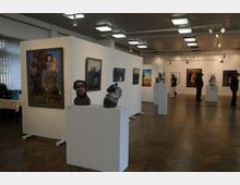 Ein Ausstellungsraum mit Holzboden, in dem Malereien und Skulpturen auf weißen Wänden und Podesten präsentiert werden. Helle Deckenbeleuchtung akzentuiert die Kunstwerke, während einzelne Besucher die Ausstellung betrachten.