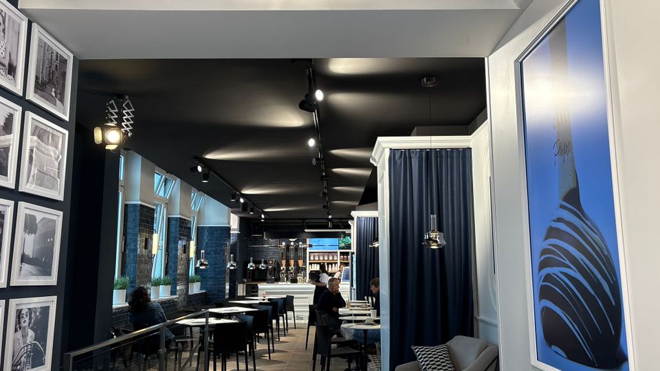 Ein modern eingerichtetes Eiscafé mit dunklem Interieur und eleganten blauen Vorhängen. Im Raum befinden sich Tische und Stühle sowie Sofas auf der rechten Seite; an der Wand hängt der Schriftzug „L’Amore per il Gelato“.
