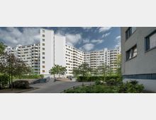 Mehrstöckige, weiße Wohngebäude mit Balkonen, die einen Innenhof umgeben, in dem sich Gehwege, Bäume und Büsche befinden. Rechts im Vordergrund ein Teil eines weiteren Gebäudes mit großen Fenstern.