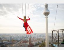 Blick von einer hohen Plattform über die Berliner Innenstadt mit dem Fernsehturm rechts im Bild. Im Vordergrund sind Stahlstreben, Seile und eine hängende Schaukel über dem Stadthorizont zu sehen.