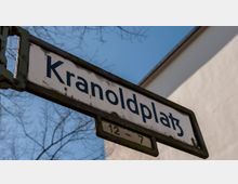 Straßenschild mit der Aufschrift „Kranoldplatz“, montiert an einer Metallhalterung, im Hintergrund ein heller Gebäudeausschnitt und kahle Baumäste vor blauem Himmel.