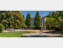 Ein Park mit Spielplatz, der aus einem Sandbereich mit Holz-Klettergerüst und einer Schaukel besteht. Im Vordergrund befinden sich eine Tischtennisplatte und ein Basketballkorb, umgeben von grünen Rasenflächen und hohen Bäumen, während im Hintergrund Wohnhäuser mit roten Ziegeldächern sichtbar sind.
