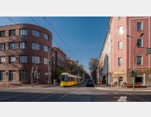 Straßenszene mit einer gelben Straßenbahn, die zwischen einem roten Backsteingebäude links und einem rosa Wohngebäude rechts entlangfährt. Die Straße ist von Bäumen gesäumt, und es sind Verkehrsampeln, Autos sowie Geschäfte im Erdgeschoss der Gebäude zu sehen.