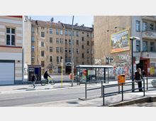 Straßenszene mit einer Bushaltestelle, einem transparenten Unterstand und einem angrenzenden Fußweg. Im Hintergrund befinden sich alte, teils verfallene Wohngebäude mit Graffiti, eine Fotoautomat-Kabine und ein Werbeplakat an einer Hauswand.