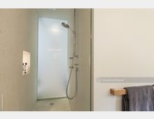 Minimalistische Dusche mit beigem Mosaikfliesenboden und -wänden, Glasabtrennung, Regendusche und Handbrause. An der Wand ist eine Nische mit Pflegeprodukten eingelassen, daneben ein Handtuchhalter mit grauem Handtuch.