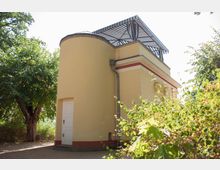 Gelbes Gebäude mit runder und eckiger Fassade, weißer Tür und einem gestreiften blauen Sonnendach auf dem Flachdach, umgeben von Bäumen und Sträuchern in einer parkähnlichen Umgebung.