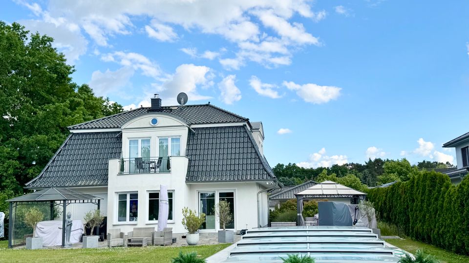 Modernes Einfamilienhaus mit weißer Fassade und dunklem Mansarddach, davor eine große Terrasse und ein langes, flaches Glasdach über einem Pool. Der Garten ist von Rasen, Hecken und Bäumen umgeben, mit Pavillons und Sitzmöbeln unter blauem Himmel mit Wolken.