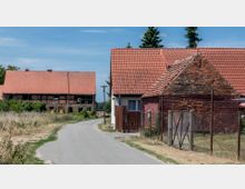 Eine ländliche Szene mit schmaler Asphaltstraße, die an mehreren Backsteingebäuden mit roten Ziegeldächern vorbeiführt. Im Hintergrund ist ein größeres Fachwerkhaus sichtbar, umgeben von Grasflächen und vereinzelten Bäumen.