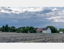 Ein ländlicher Acker mit einer einfachen, weiß-grauen Scheune mit rotem Dach am rechten Bildrand, umgeben von Bäumen und dichtem Grün unter einem bewölkten Himmel.