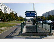 U-Bahn-Eingang mit dem Schild „Strausberger Platz“ in einer urbanen Umgebung, flankiert von Grünflächen sowie mehrstöckigen Wohngebäuden und einer Straße mit parkenden Autos und Bäumen. Im Hintergrund ist eine klassische Uhr an einem Mast zu sehen.
