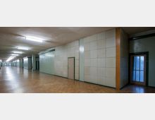 Langer, rechteckiger Flur mit hellbraunem Parkettboden, grünlichen Wänden und einer Tür mit Glasfenstern auf der rechten Seite. Deckenleuchten sorgen für gleichmäßige Beleuchtung, und der Gang ist in verschiedene Abschnitte durch Säulen unterteilt.