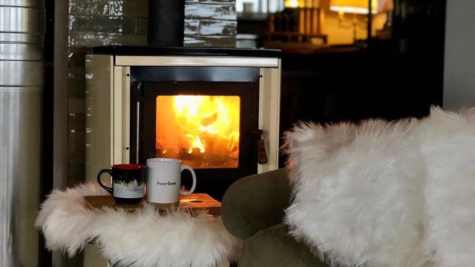Gemütliche Inneneinrichtung mit einem brennenden Kaminofen vor einer glänzenden, dunklen Fliesenwand. Ein grüner Sessel mit weißem Fellüberwurf und ein kleiner Tisch mit Fellauflage und Tassendekoration stehen im Vordergrund, während eine warme Beleuchtung den Hintergrund erhellt.