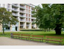 Mehrstöckiges modernes Wohngebäude mit Balkonen, umgeben von einer Grünanlage mit gepflegtem Rasen, großen Bäumen und mehreren Holzbänken auf einem Kiesweg im Vordergrund.