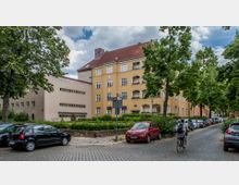 Mehrgeschossiges Wohnhaus mit gelber Fassade und rot gedecktem Dach in einer von Bäumen gesäumten Straße. Parkende Autos sowie ein Fahrradfahrer auf Kopfsteinpflaster prägen das Straßenbild, im Hintergrund ein weiteres Gebäude mit heller Putzfassade.