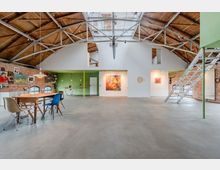 Großer, loftartiger Innenraum in einer umgebauten Industriehalle mit sichtbaren Stahlträgern, hohem Holztonnen­dach und Backsteinwänden. Offene Fläche mit Esstisch und Stühlen, kleiner Küchenzeile, Wandkunst sowie einer Metalltreppe zu einer oberen Galerieebene.
