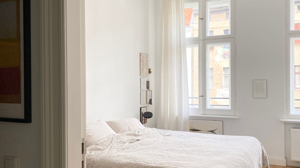 Helles Schlafzimmer mit weißem Doppelbett und hohen, weiß gerahmten Fenstern mit transparenten Vorhängen. Weiße Wände mit wenigen Bildern, eine schwarze Deckenlampe und ein heller Holzboden mit Teppich prägen den Raum.