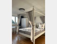 Ein Schlafzimmer mit einem weißen Himmelbett, das graue Vorhänge und Bettwäsche mit Kirschmuster hat. Holzboden, ein Fenster mit Tageslicht, ein weißer Kleiderschrank und ein kleiner runder Tisch ergänzen die Einrichtung.