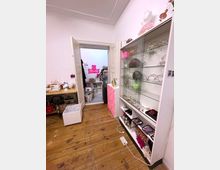 Ein kleiner Innenraum mit Holzboden, Regalen mit Dekorationsartikeln und Schmuck auf der rechten Seite sowie einem offenen Durchgang zu einem angrenzenden Arbeitsraum. Im Hintergrund sind ein Schreibtisch, mehrere Kartons und Neon-Wandschilder sichtbar.