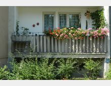 Ein Balkon an einem Wohngebäude mit wellenförmiger Metallverkleidung, dekoriert mit blühenden rosa und roten Blumen, sowie kletternden Pflanzen. Vor dem Balkon befinden sich Sträucher und eine kleine Leiter lehnt an der Verkleidung.