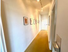 Langer, schmaler Innenflur mit weißen Wänden und beigem Teppichboden, an der linken Wand mehrere gerahmte Bilder. An der Decke sind Spotleuchten montiert, am Ende des Gangs befindet sich eine weiße Tür.