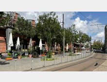 Straßenszene mit einer gepflasterten Fläche und einer Straßenbahnschiene im Vordergrund. Im Hintergrund steht ein Backsteingebäude mit Bäumen, Außensitzbereichen und Sonnenschirmen, die zu Cafés oder Restaurants gehören.