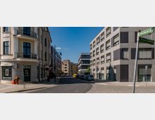 Straßenkreuzung in einer Stadt mit einem historischen Gebäude auf der linken Seite, das helle Fassaden und Balkone mit geschmiedeten Geländern hat, und einem modernen Bürogebäude mit grauer Fassade und rechteckigen Fenstern auf der rechten Seite. Im Hintergrund sind weitere Gebäude und geparkte Autos zu sehen, bei blauem Himmel und ruhiger Atmosphäre.