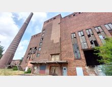 Backstein-Gebäude eines verlassenen Industriegeländes mit zerbrochenen Fenstern und einer großen Schornsteinstruktur im Hintergrund. Im Vordergrund sind verwitterte Tore, bröckelnder Putz und ein bewachsener Zaunbereich sichtbar.