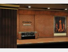 U-Bahn-Station mit Backsteinwänden, der Schriftzug „Zitadelle“ und ein Ausgangswegweiser sind sichtbar. Ein Wandgemälde eines historischen Porträts und schwarze Sitzbänke ergänzen die Gestaltung.