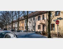 Eine Reihe von zweistöckigen Reihenhäusern mit pastellfarbenen Fassaden, umgeben von kahlen Bäumen entlang einer Straße. Im Vordergrund sind parkende Autos und ein Straßenschild mit einem Durchfahrtverbot zu sehen.