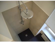 Duschbereich in einem Badezimmer mit beigen Wandfliesen, einer runden, verchromten Regendusche und einem Boden aus kleinen, dunklen Fliesen mit einem zentralen Abfluss. Rechts daneben befindet sich ein Fenster und ein Heizungskörper.