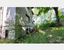 Eine Seitenansicht eines mehrstöckigen Wohngebäudes mit Putzfassade; vor dem Haus führt eine Treppe durch einen überwucherten Garten mit hohen Bäumen und dichtem Grün. Die Umgebung wirkt schattig und von natürlicher Vegetation geprägt.