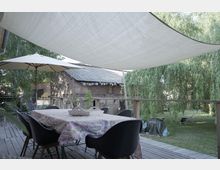 Eine Terrasse mit Holzboden und einem großen Tisch, der mit einer gemusterten Tischdecke bedeckt ist, umgeben von schwarzen Stühlen. Darüber spannen Sonnensegel, während im Hintergrund ein Backsteingebäude und ein Garten mit Weidenbäumen und Liegestühlen zu sehen sind.